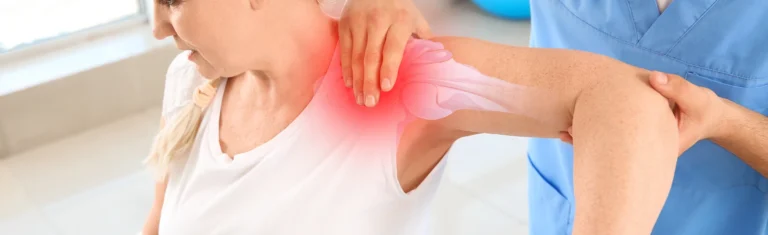 A physiotherapist helps a woman stretch her shoulder. An overlay shows the shoulder joint and a red area highlights pain or injury. The woman wears white; the therapist wears blue scrubs.