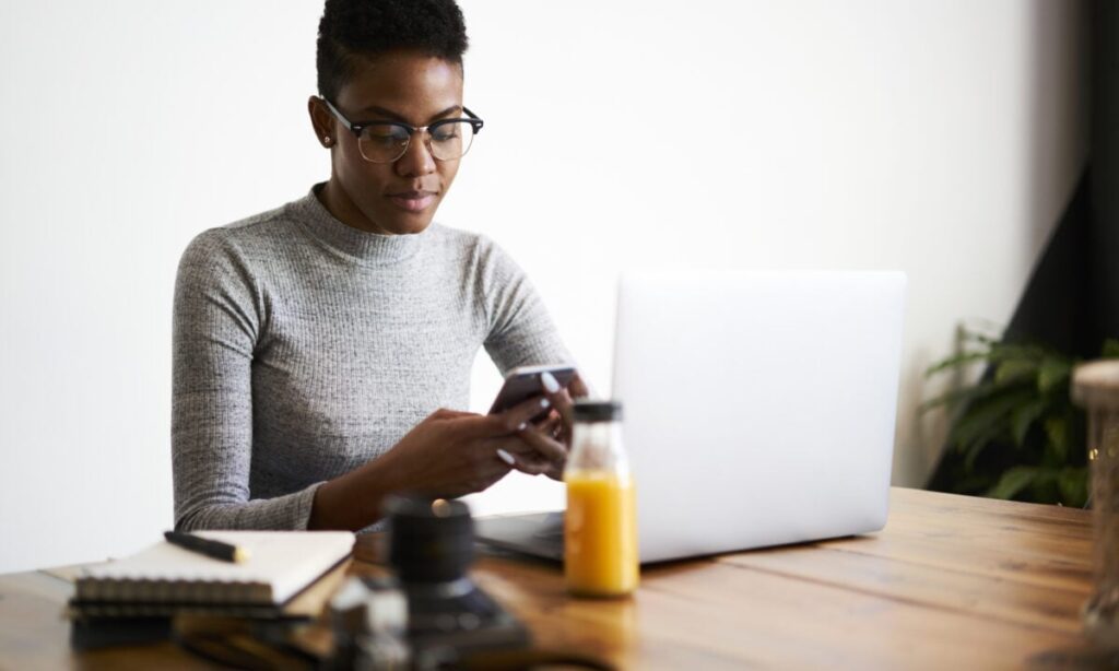 A person wearing glasses and a grey jumper sits at a wooden table, looking at their phone. A laptop, notebook, camera lens, and a bottle of orange juice are on the table. A plant is visible in the background.