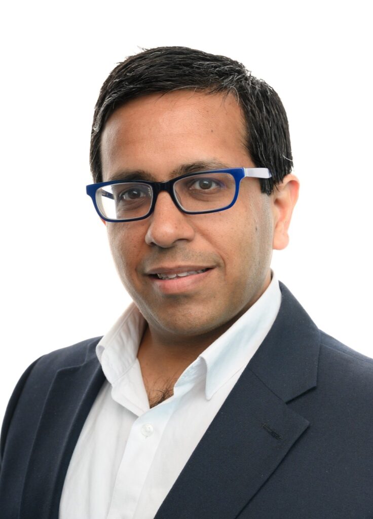 A man with short dark hair, wearing blue-rimmed glasses, a navy blazer, and a white collared shirt, poses against a plain white background.