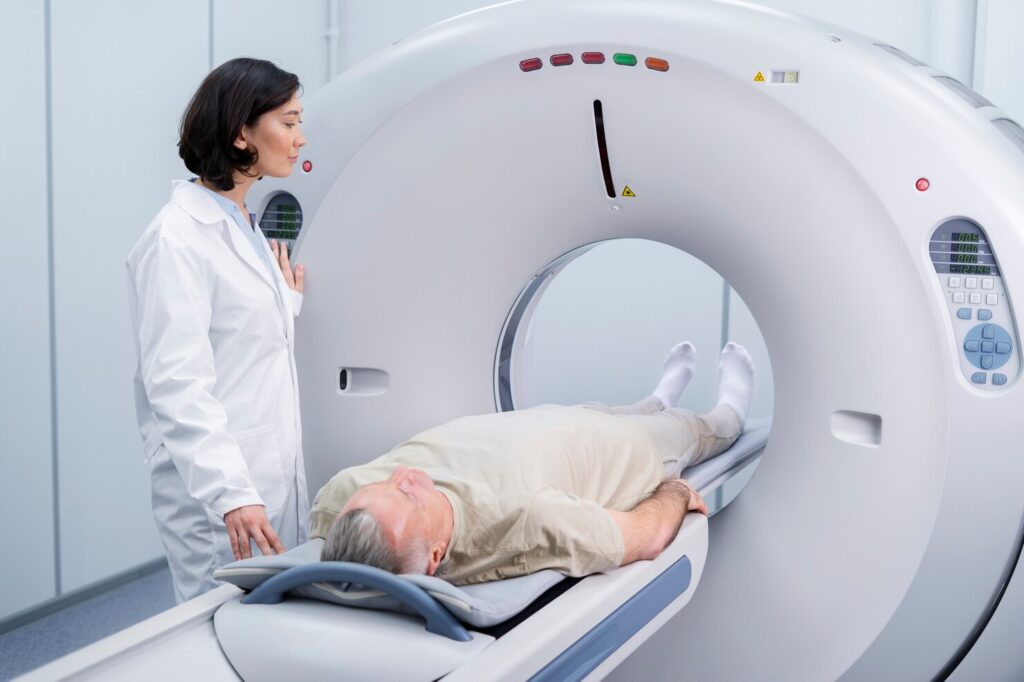 A medical professional stands beside an older man lying on a bed, entering a CT scan machine for a medical imaging procedure in a clinical setting.