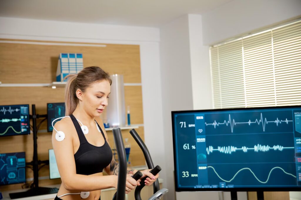 A woman wearing fitness sensors exercises on a machine in a lab. Medical monitors display her heart rate and other vital signs, with graphs visible on large screens. The room is bright, with shelves and blinds in the background.