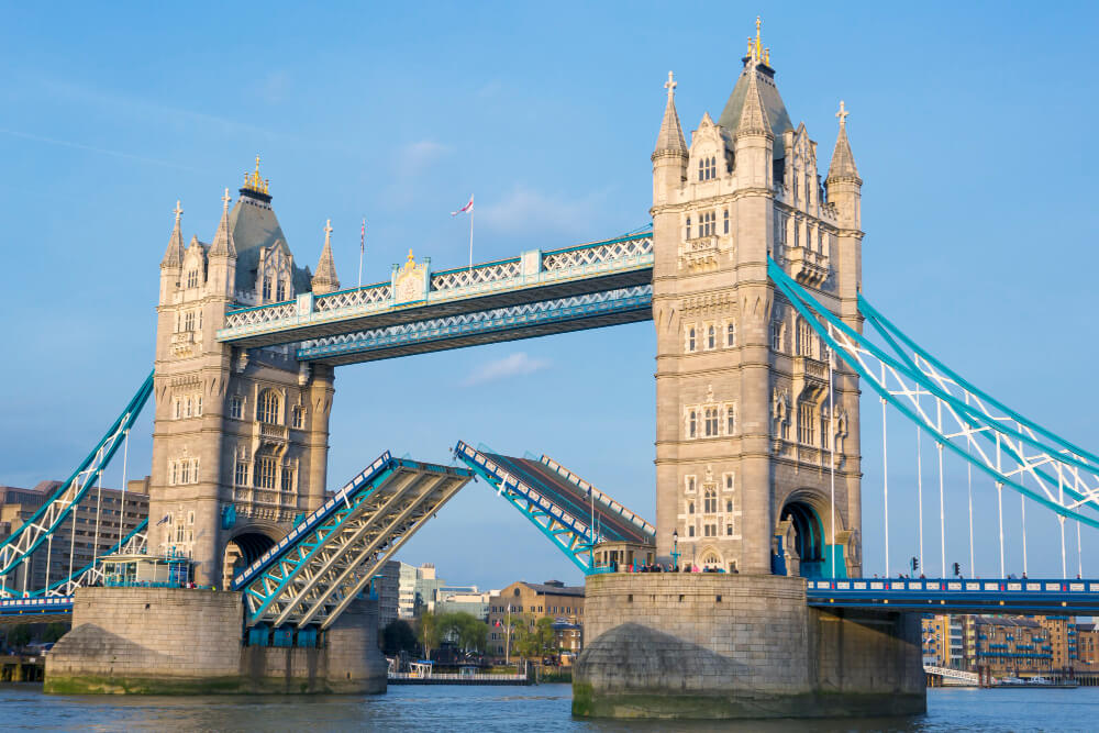 The photo shows London’s Tower Bridge with its two tall towers and blue suspension sections. The bridge’s roadway is raised, allowing boats to pass underneath on the River Thames. The sky is clear and blue.