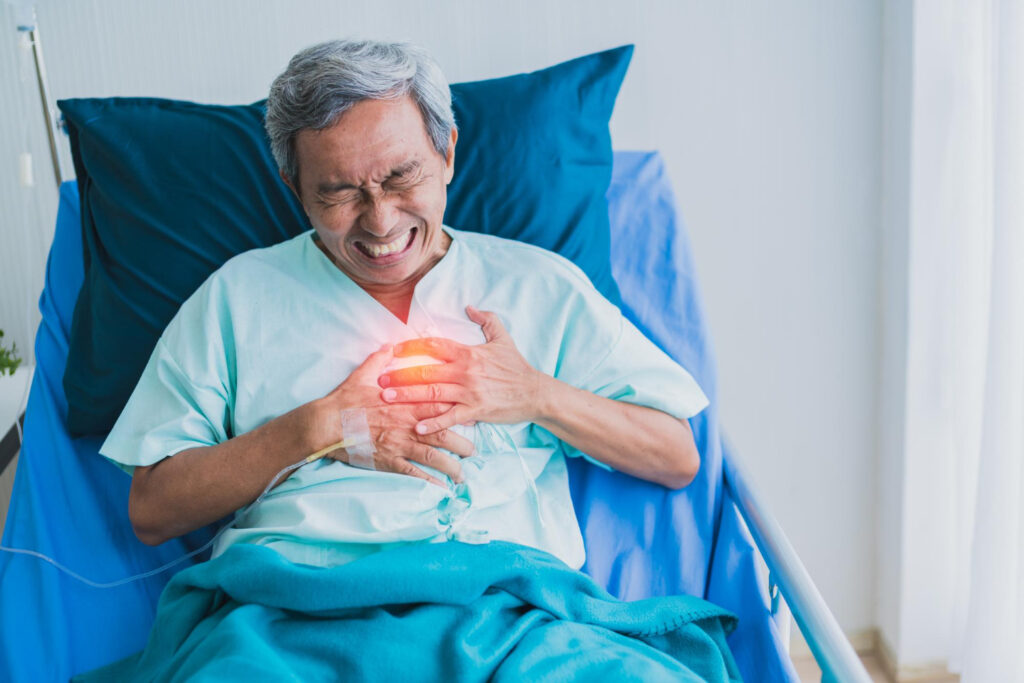 An older man in a hospital bed clutches his chest with a pained expression, suggesting severe chest pain. A glowing red spot highlights the chest area, indicating a possible heart problem or cardiac event.