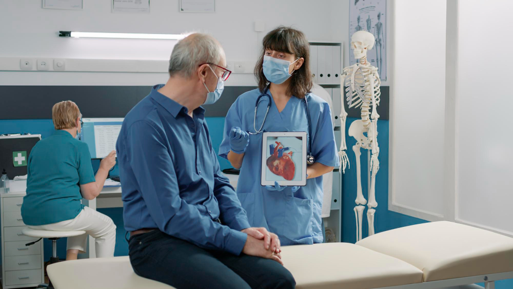 A doctor wearing a mask shows a heart diagram to an older male patient sitting on an examination table, while another healthcare worker types at a computer in the background. A skeleton model stands nearby.