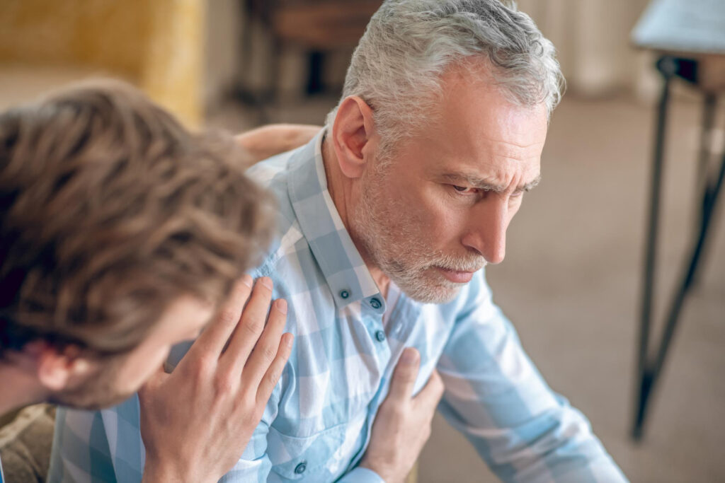 A younger man comforts an older man with grey hair and a checked shirt, gently placing a hand on his shoulder as the older man looks distressed or deep in thought.