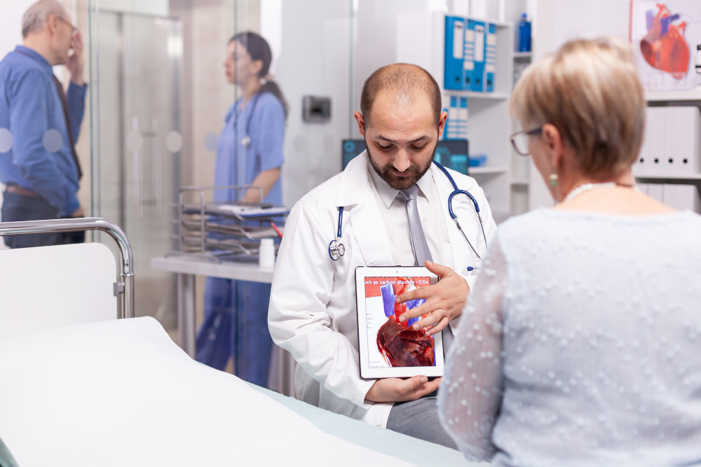 A doctor in a white coat shows a heart diagram to a seated patient in a medical surgery. In the background, two people, one in scrubs, talk near a glass wall. Medical equipment and charts are visible.