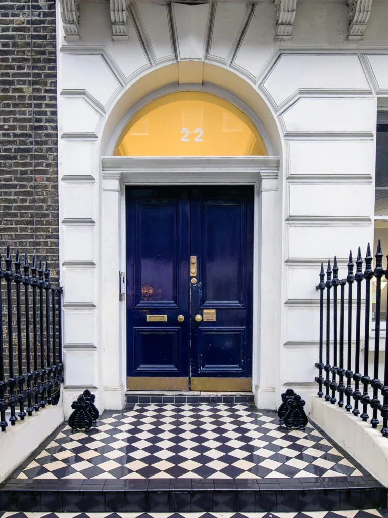 A dark blue double front door with gold fittings, set in a white arched doorway with the number 22 above. The entrance features black-and-white chequered tiles and black iron railings on each side.