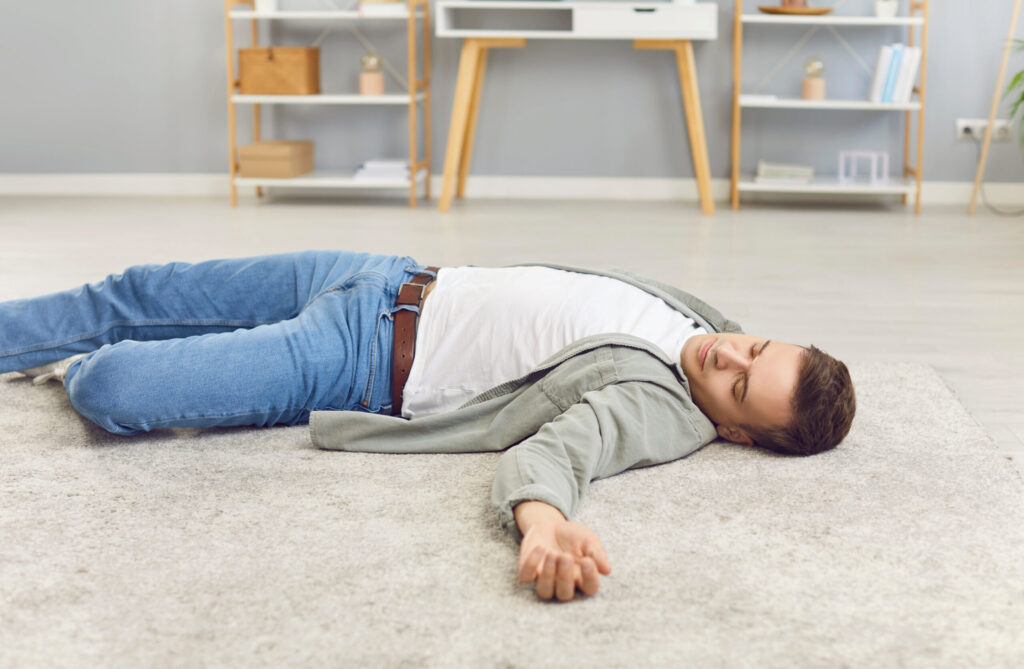 A man in casual clothes lies on his back on a carpeted floor in a bright living room, eyes closed and arm extended, appearing unresponsive or resting. Shelves and a desk are visible in the background.